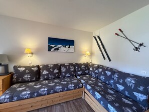 A cozy room with a sectional sofa, two wall-mounted lamps, a painting of a snowy landscape, and skis and ski poles on the wall.