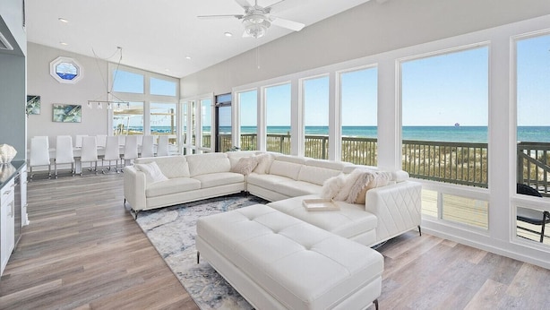 A spacious living room with a large white sectional sofa, a ceiling fan, and a view of the ocean through large windows. - Beachfront Bliss: Spacious Deck, Grill & Unmatched Gulf Views! (Panama City Beach)