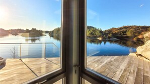 Vista desde una habitación con un ventanal grande que da a un río tranquilo, una terraza de madera y un promontorio rocoso.