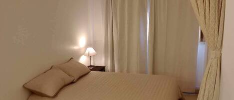 Un dormitorio con una cama, dos almohadas, una lámpara de noche y una ventana con cortinas.