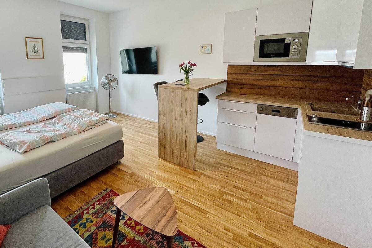 Eine kompakte, moderne Wohnung mit Bett, einer kleinen Couch, einem Holztisch, einer Küche mit weißen Schränken und einer Mikrowelle sowie einem an der Wand montierten Fernseher.