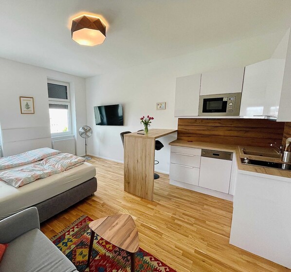 A compact, modern apartment with a bed, a small sofa, a wooden coffee table, a kitchen with white cabinets and a microwave, and a TV mounted on the wall.