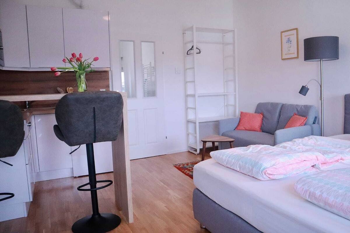 Ein modernes, gut beleuchtetes Zimmer mit einem Bett, einem grauen Sofa, einem kleinen Tisch und einem Stuhl. Es gibt einen weißen Kleiderschrank und eine Kleiderstange mit Kleidung.
