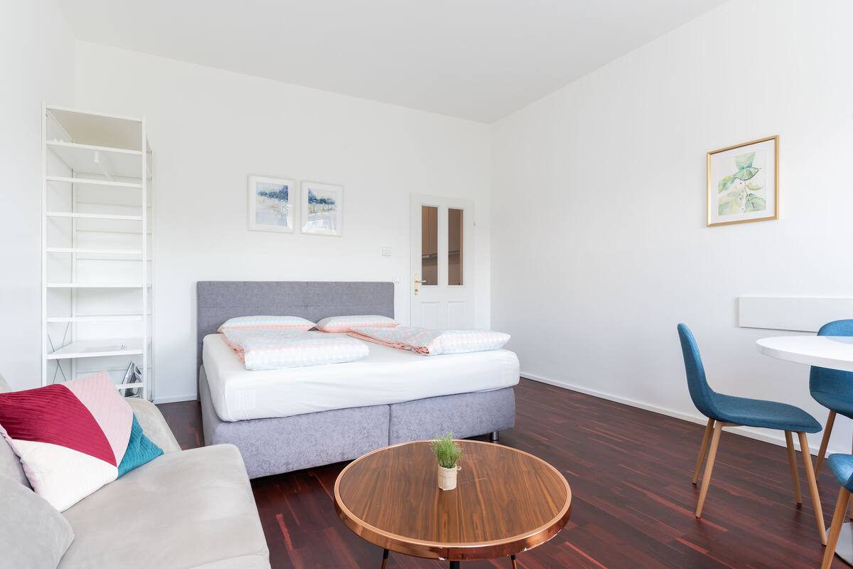 Ein modernes Schlafzimmer mit einem Bett, einem runden Holztisch, einem grauen Sofa und einem Essbereich mit Stühlen.