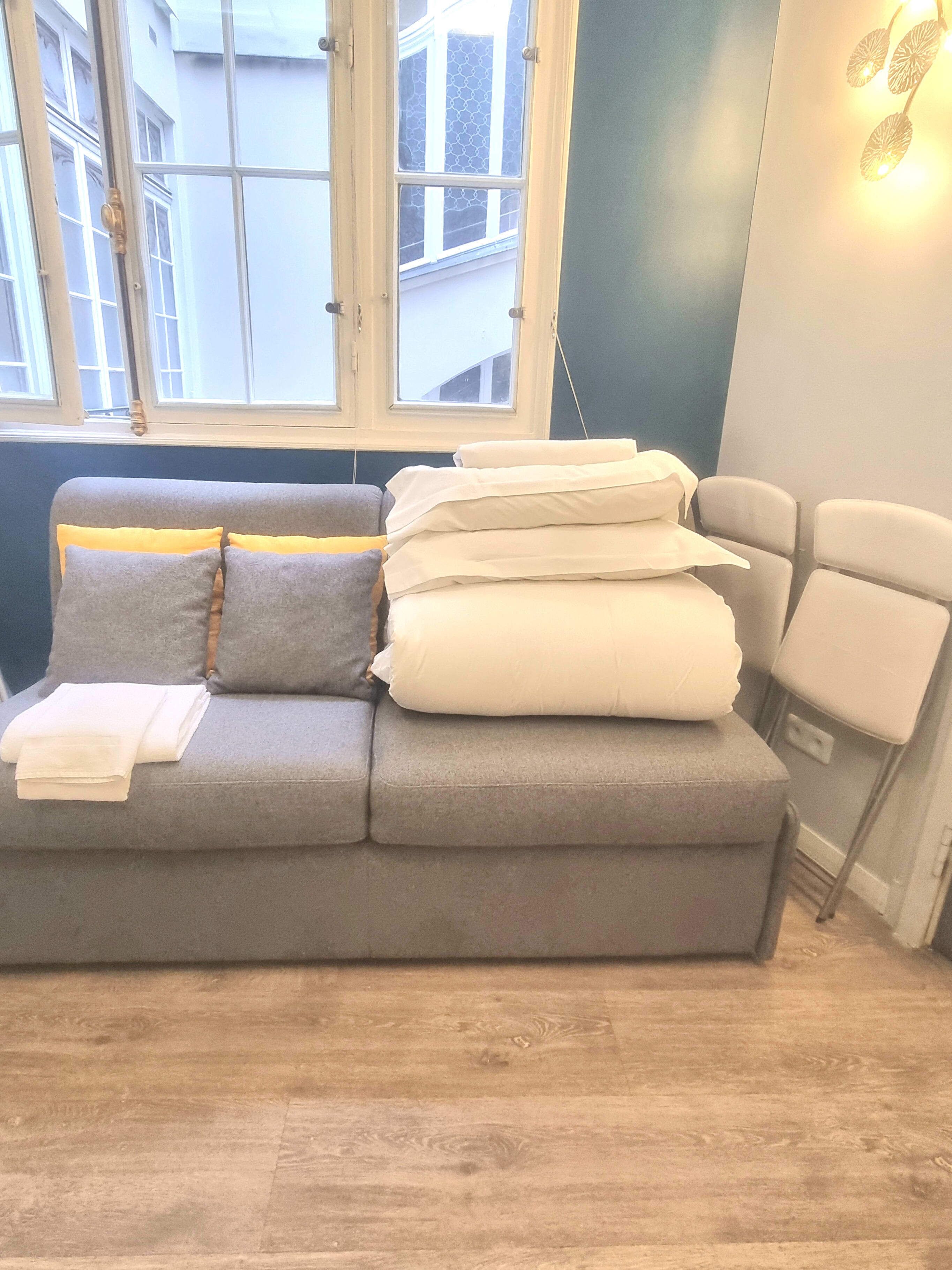 A grey sofa with cushions and folded blankets, a window with a view of buildings, and a wall-mounted light fixture.
