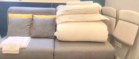 A grey sofa with cushions and folded blankets, a window with a view of buildings, and a wall-mounted light fixture.