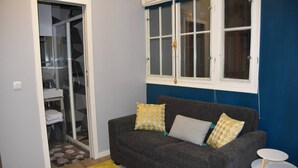 A room with a grey sofa, two white chairs, a glass shower door, a window with white frames, and a blue wall.