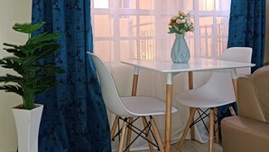A dining area with a table and chairs, a potted plant, and a window with blue curtains. - Lux Suites Neema court Apartments (Nairobi)