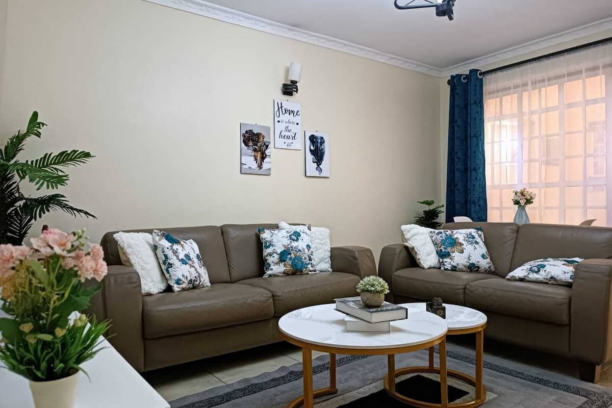 A living room with two brown sofas, a white coffee table, and decorative pillows. There are wall art pieces and a potted plant.