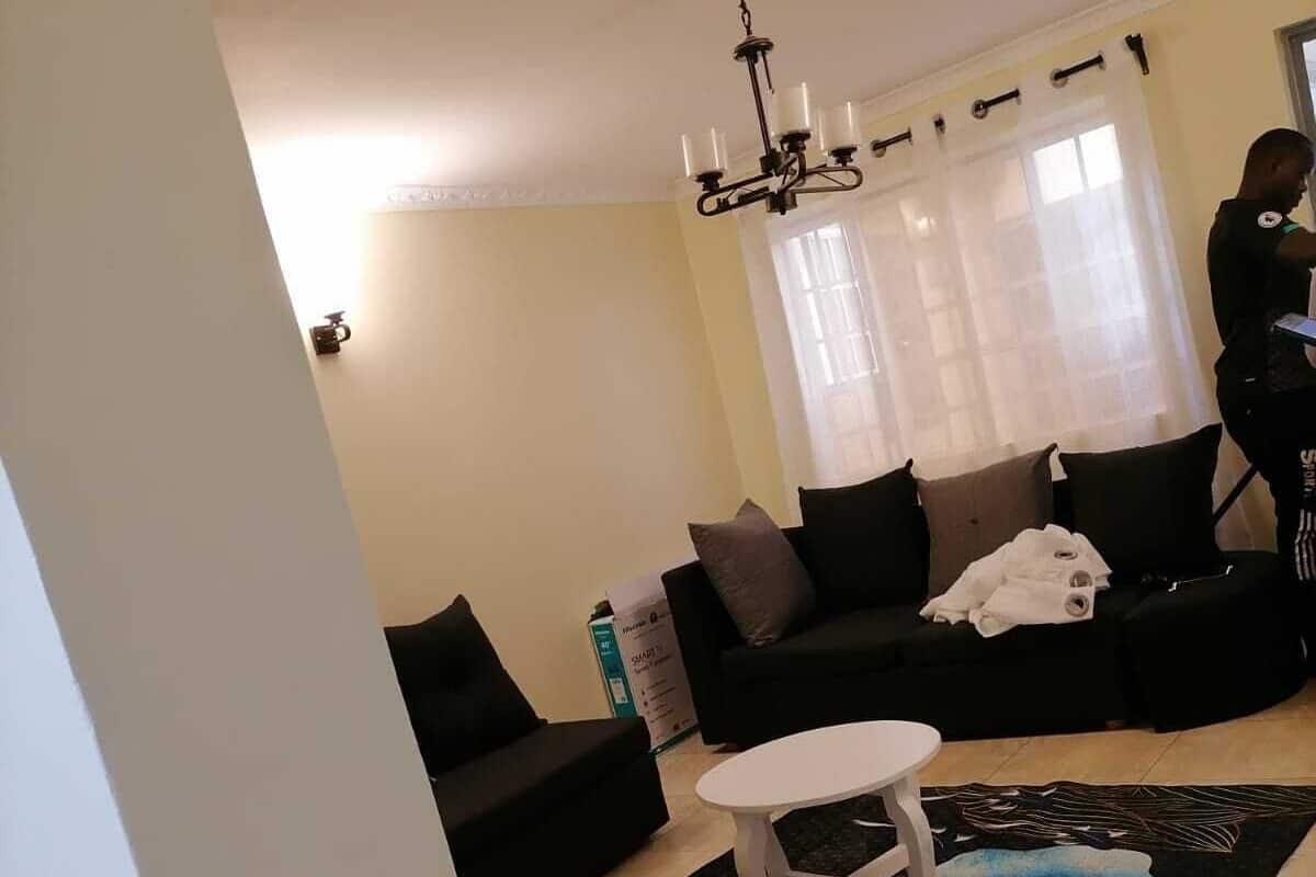 A living room with a black sofa, a white coffee table, a white side table, a chandelier, and a window with sheer curtains.