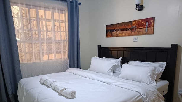A neatly made bed with white linens and a rolled towel, a dark wooden headboard, and a window with blue curtains. - Lux Suites Neema court Apartments (Nairobi)
