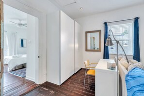 A modern bedroom with a bed, a desk, and a chair. There is a window with curtains and a ceiling fan. - Irish Channel Apt w/ Balcony | Walk to Magazine St (New Orleans)