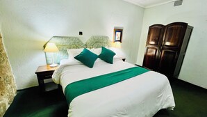 A neatly made bed with a green bedspread and two pillows, a nightstand with a lamp, and a wooden wardrobe.