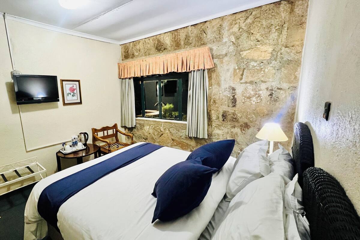 A hotel room with a bed, a television, a small table with a tray of items, a window with curtains, and a stone wall.