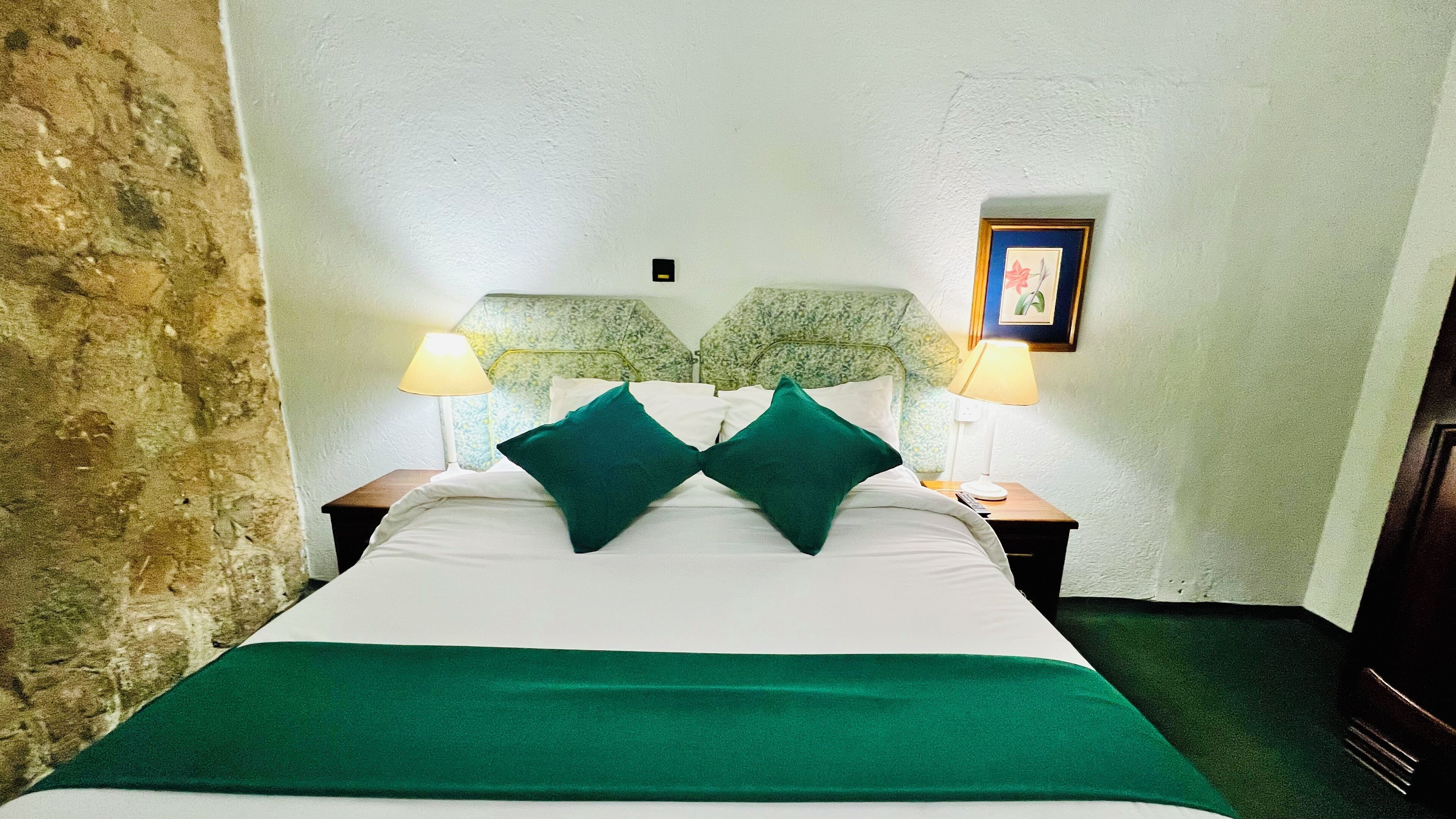 A bed with a green bedspread and two green pillows, flanked by two nightstands with lamps, in a room with a stone wall and a framed picture on the wall.