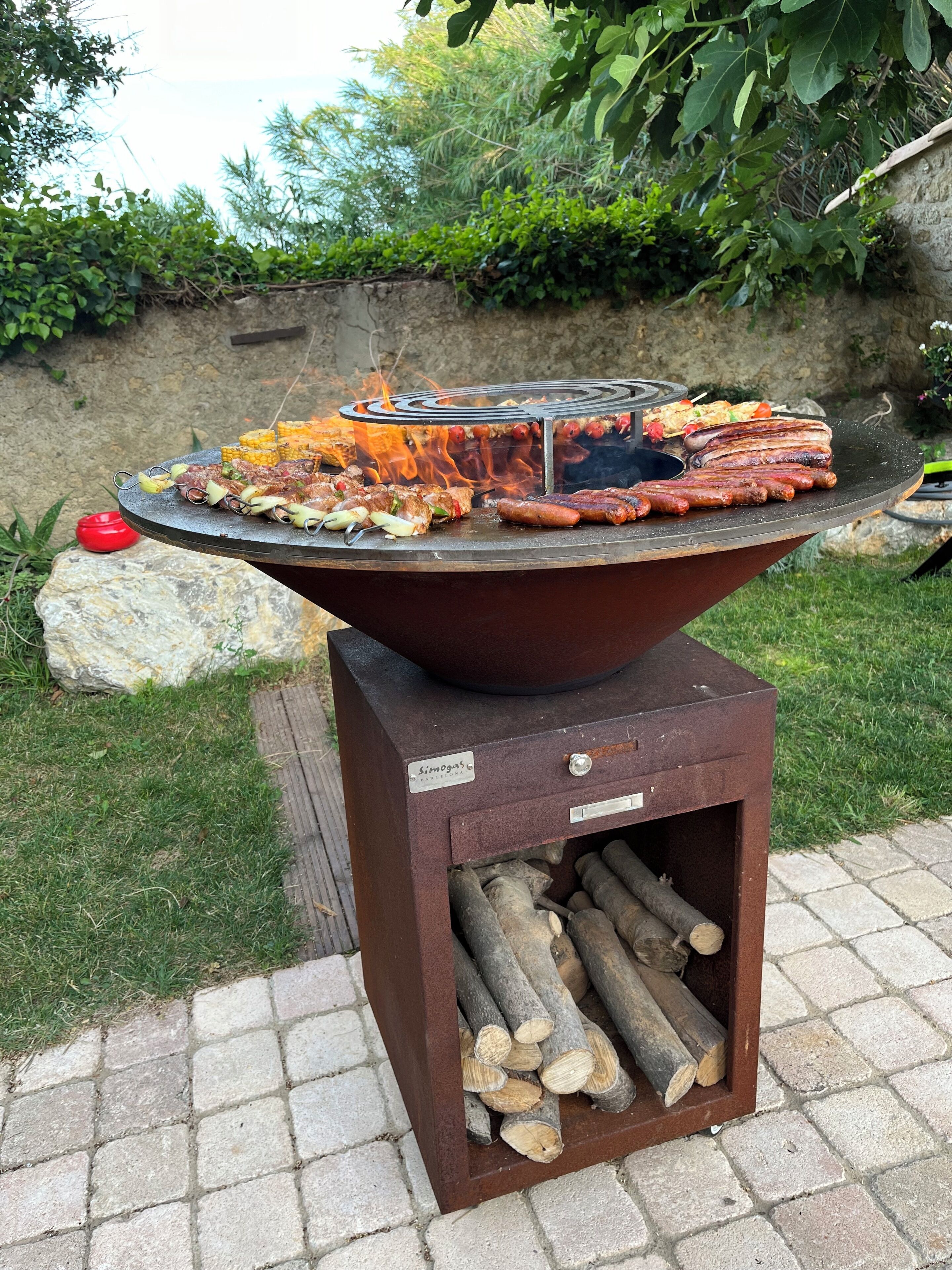 A round outdoor grill with various meats cooking, surrounded by a stone wall and greenery.