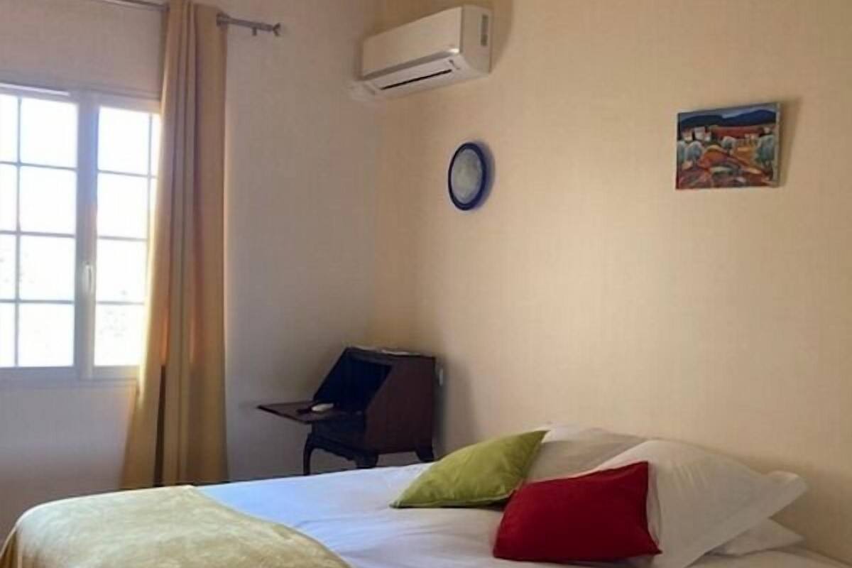A hotel room with a bed, a chair, an air conditioning unit, a clock, and a painting on the wall.