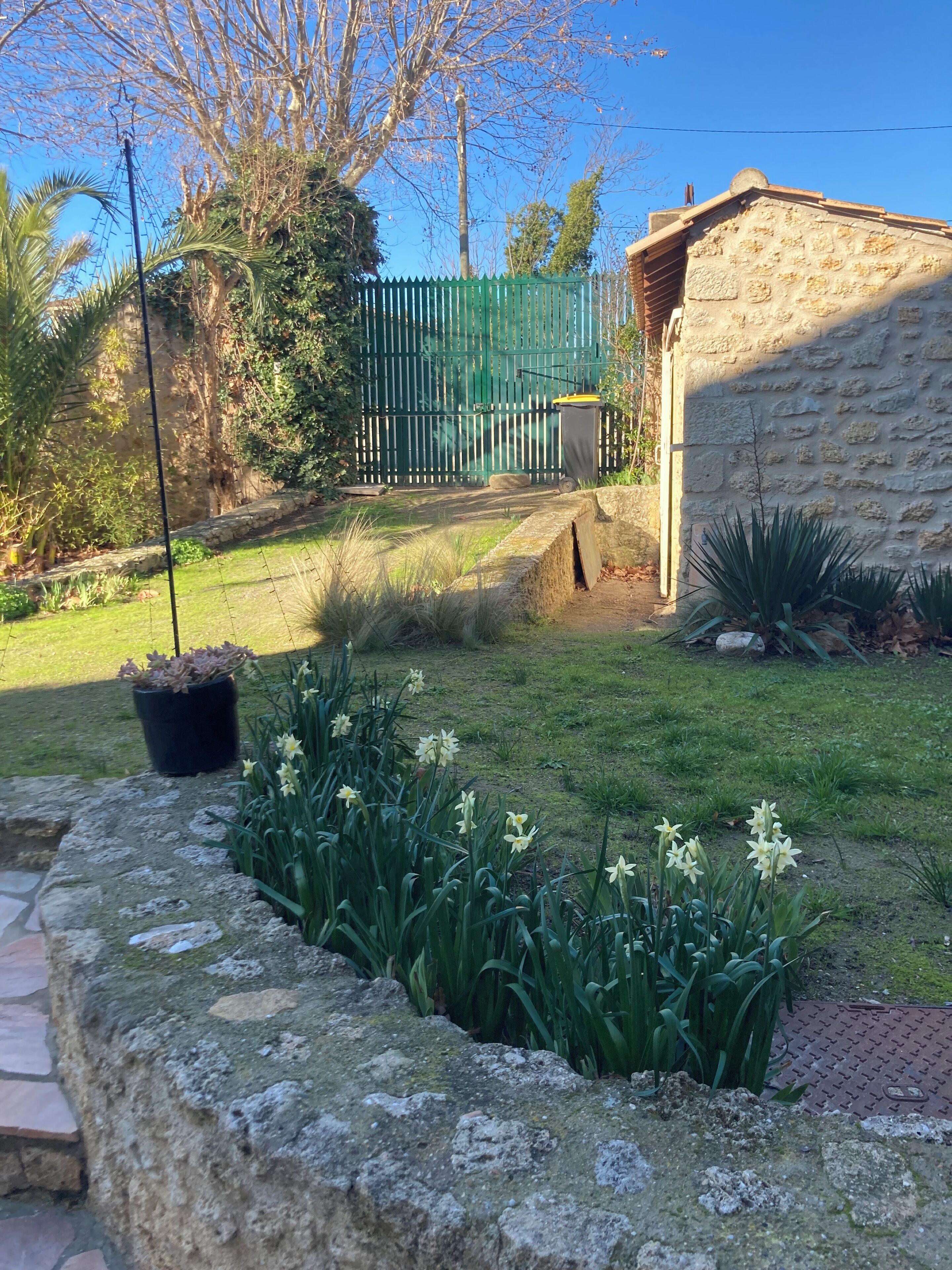 Un edificio de piedra con una cerca verde, un camino de piedra y plantas con flores.