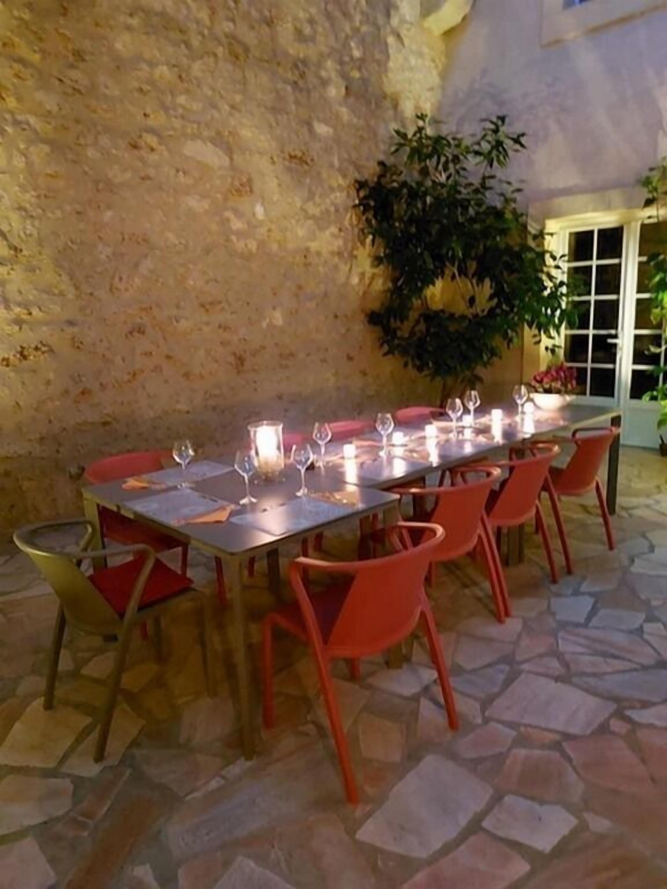An outdoor dining area with a stone floor, a long table set with wine glasses and candles, surrounded by orange chairs.