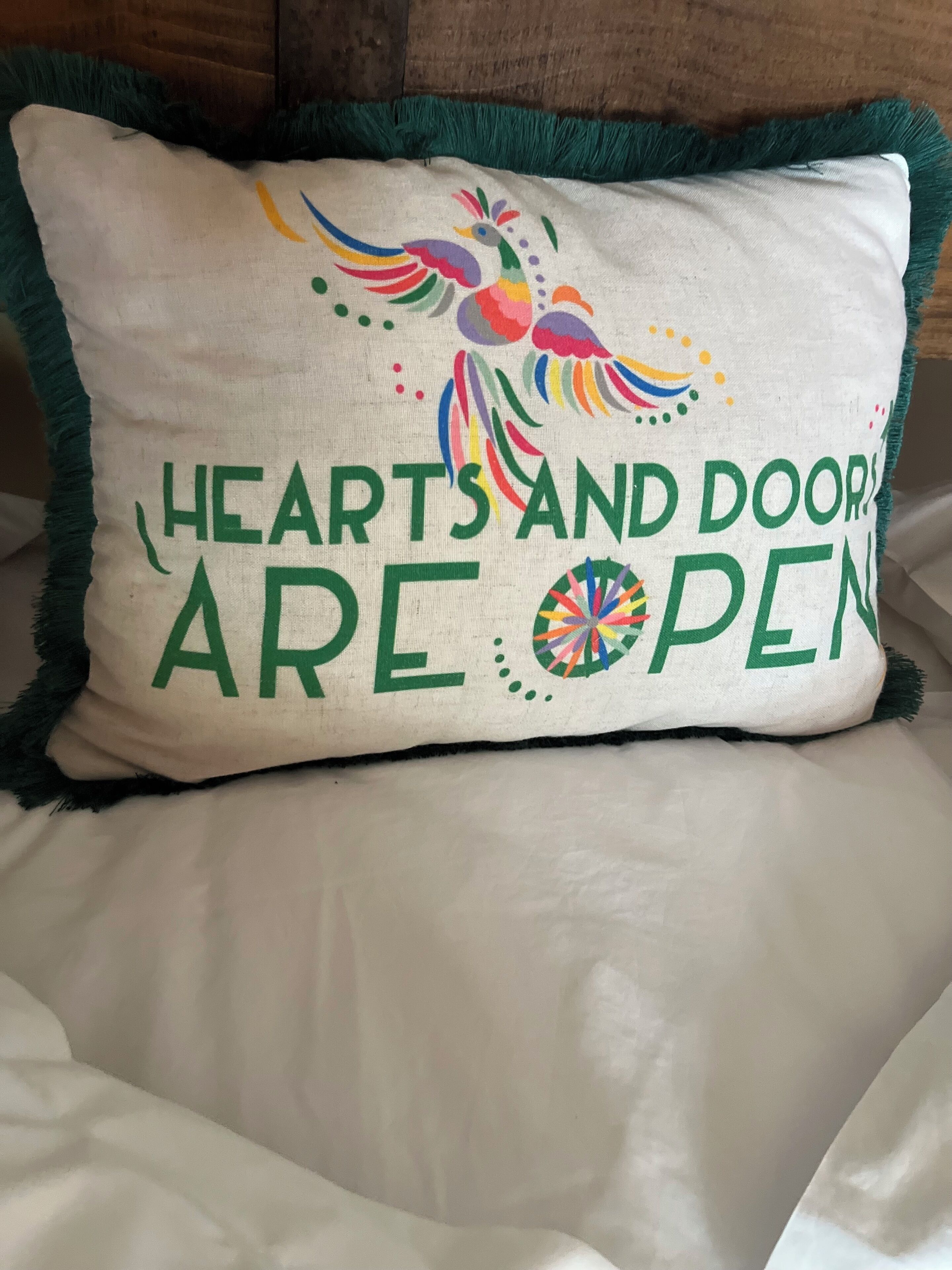 Un coussin orné du texte « Les cœurs et les portes sont ouverts » et d’un motif représentant un oiseau aux couleurs vives.
