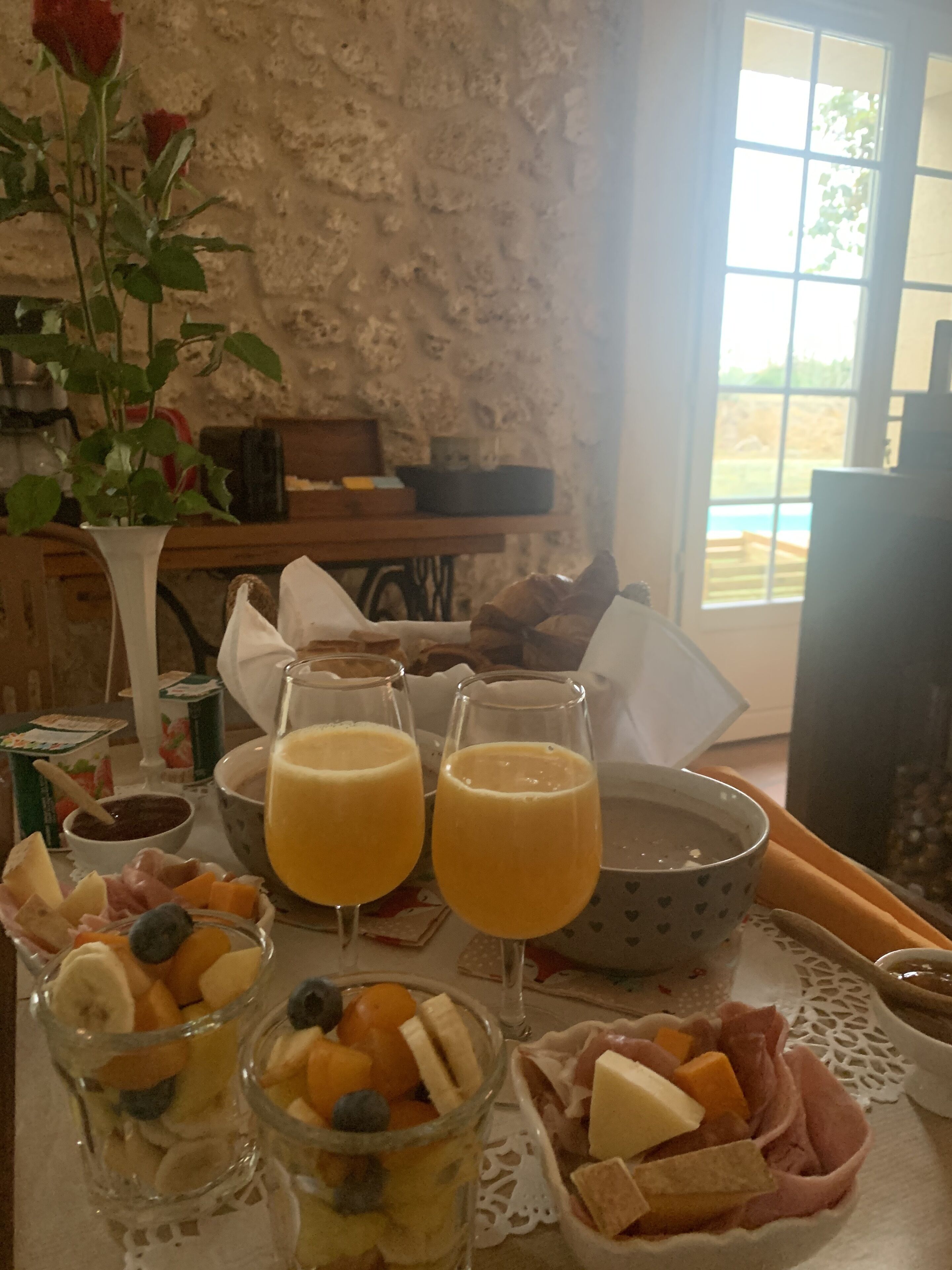 A breakfast table with fruit, cheese, and drinks, a vase with flowers, and a rustic stone wall.