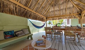 Un espacio de estar al aire libre con techo, una hamaca, muebles de madera y una mesa con un libro y un vaso.