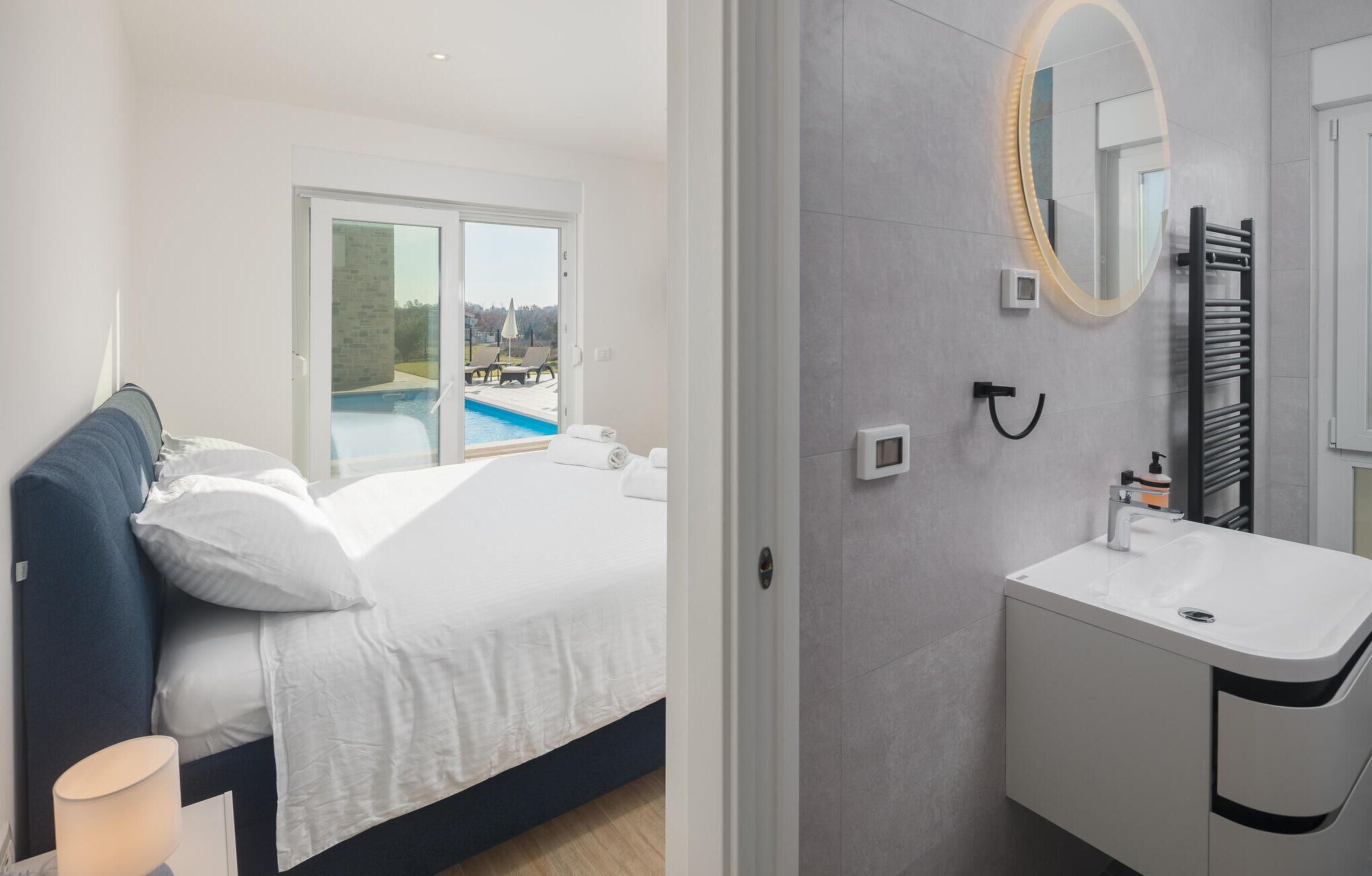 A modern hotel room with a bed, a bathroom with a sink and mirror, and a view of a swimming pool through the window.