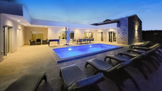 A modern outdoor pool area with lounge chairs, a dining table, and a stone wall. - Beach front home in Babici (Babici)