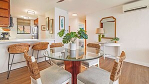A dining area with a glass table, wicker chairs, a ceiling fan, and a kitchen in the background. - Top Floor Condo- Ocean Views w Pool Steps to Beach (Wailuku)