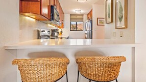 A kitchen with wooden cabinets, a white countertop, two woven chairs, and framed botanical prints on the wall. - Top Floor Condo- Ocean Views w Pool Steps to Beach (Wailuku)
