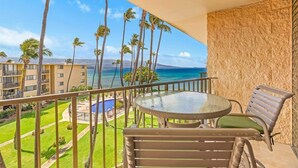 A balcony with a round glass table and two chairs, overlooking a pool and palm trees. - Top Floor Condo- Ocean Views w Pool Steps to Beach (Wailuku)