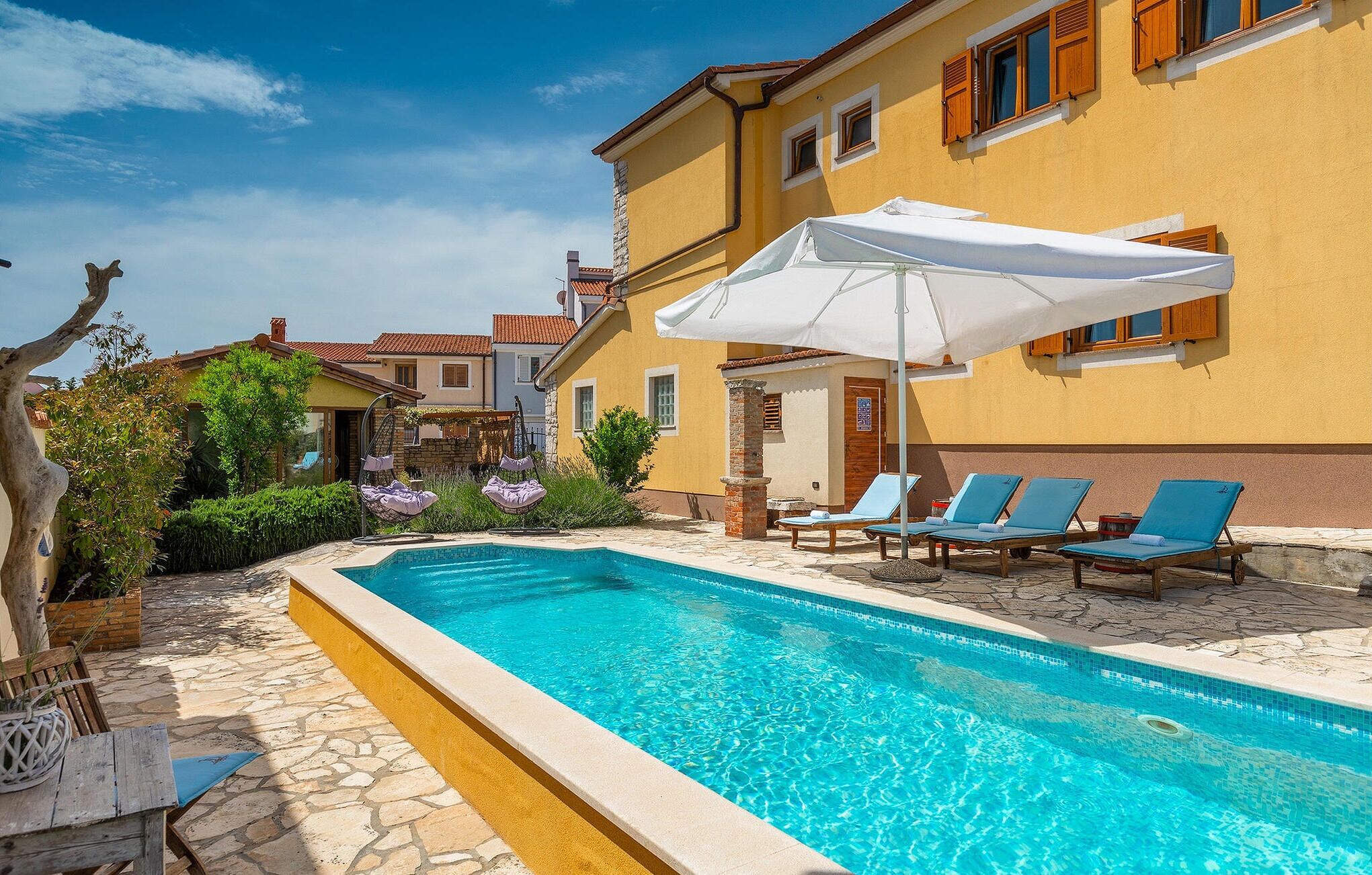 Una piscina con sillas de descanso y sombrillas, rodeada de suelo de piedra y un edificio amarillo con contraventanas de madera.