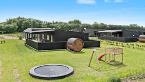 Ein modernes Gebäude mit einem großen Holzbottich, einem Trampolin und einer Schaukel auf einer grünen Wiese.