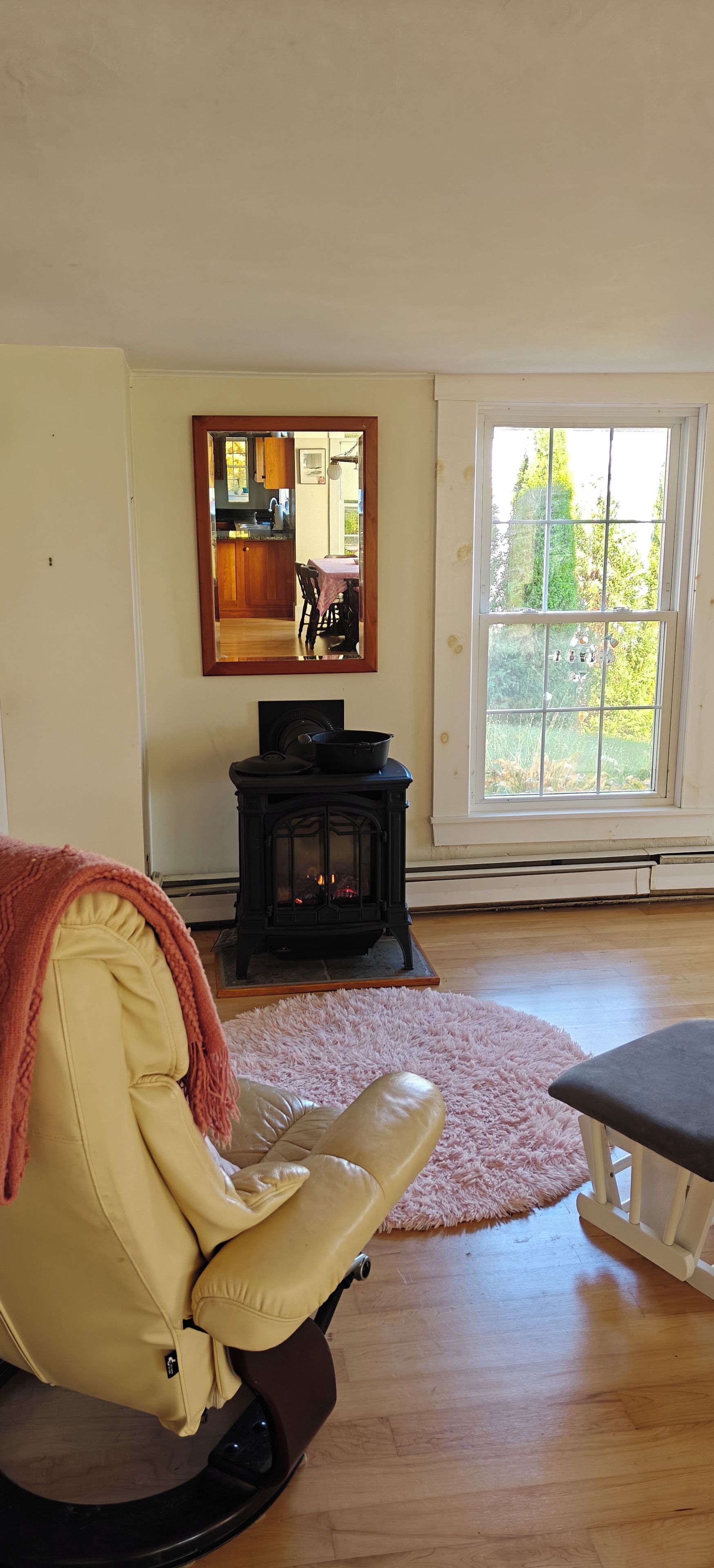 A room with a leather chair, a table, a fireplace, a window with a view of greenery, and a mirror on the wall.