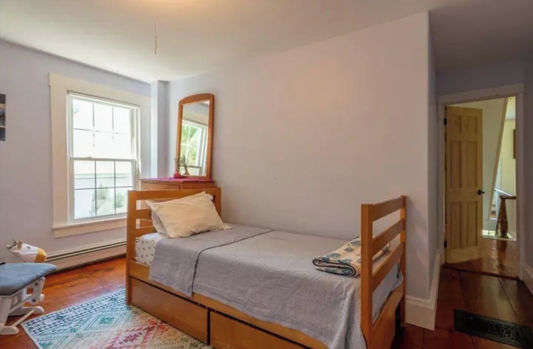 A single bed with a headboard, a bedside table, a mirror, and a window with blinds.