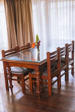A dining table with glass top and wooden chairs, set in a room with wooden flooring and curtains. - ELIXIR WOODS RESORT AND SPA (Devikolam)