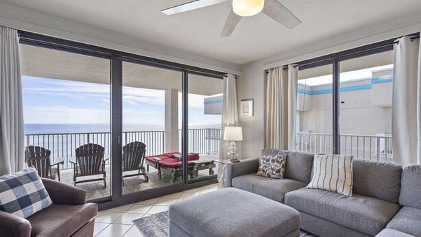 A modern living room with a grey sofa, ottoman, and armchair, a glass coffee table, and a view of the ocean through large sliding glass doors. - Windward Pointe 1206 by Liquid Life (Orange Beach)