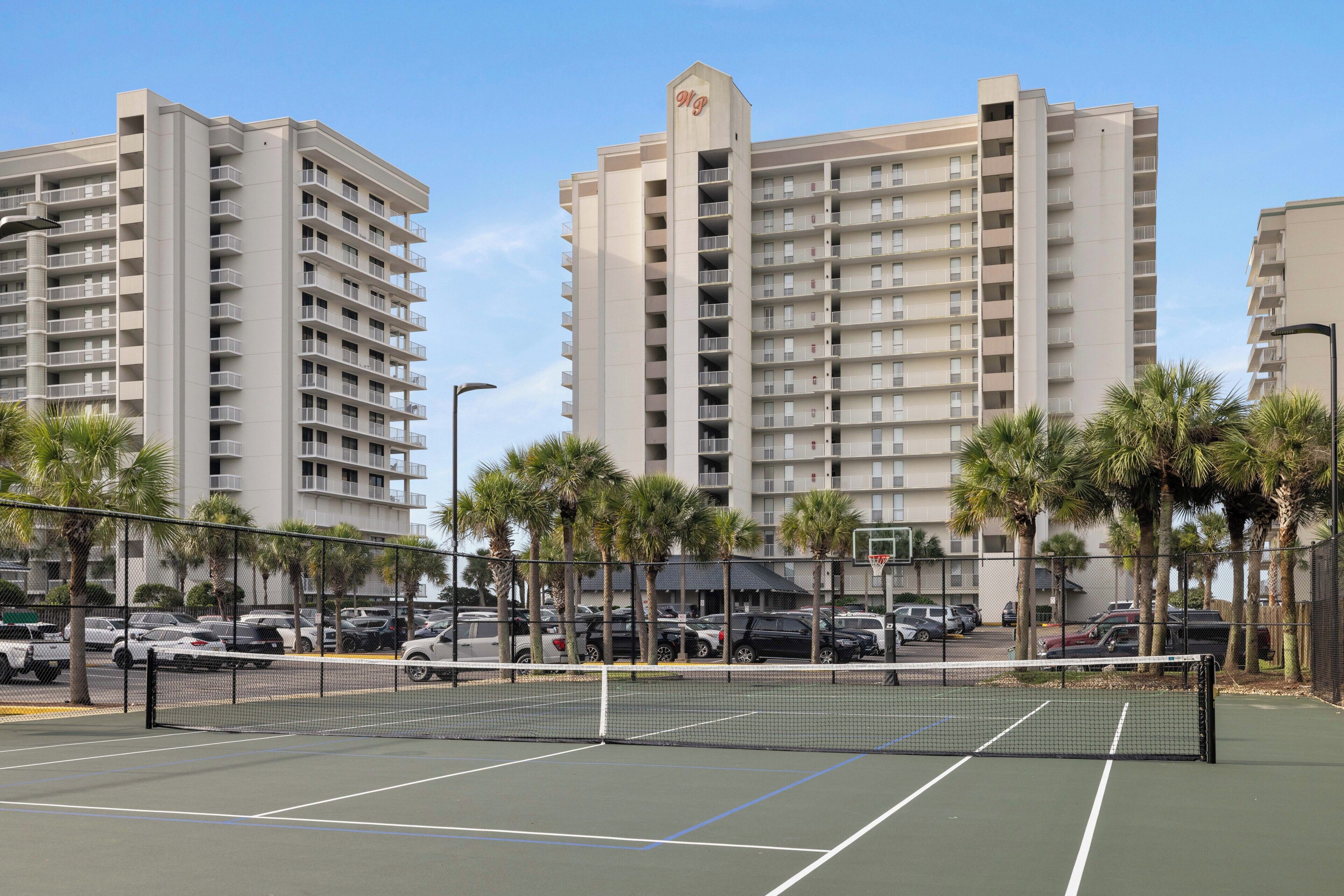 A tennis court with a net, surrounded by a fence, palm trees, and a parking lot with cars.