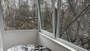 A corner room with a round table and a cushioned chair, a large window with a view of leafless trees, and a white wall. - Apartament la 3 minute de aeroport, in centrul orasului langa parc si restaurant (Otopeni)