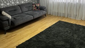 A living room with a grey sofa, a black rug, wooden flooring, floral wallpaper, and sheer curtains. - Apartament la 3 minute de aeroport, in centrul orasului langa parc si restaurant (Otopeni)