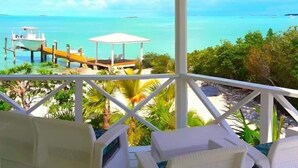 Un espace extérieur aménagé, abrité et doté de mobilier en osier blanc, offrant une vue sur une plage avec un quai et un petit bâtiment.