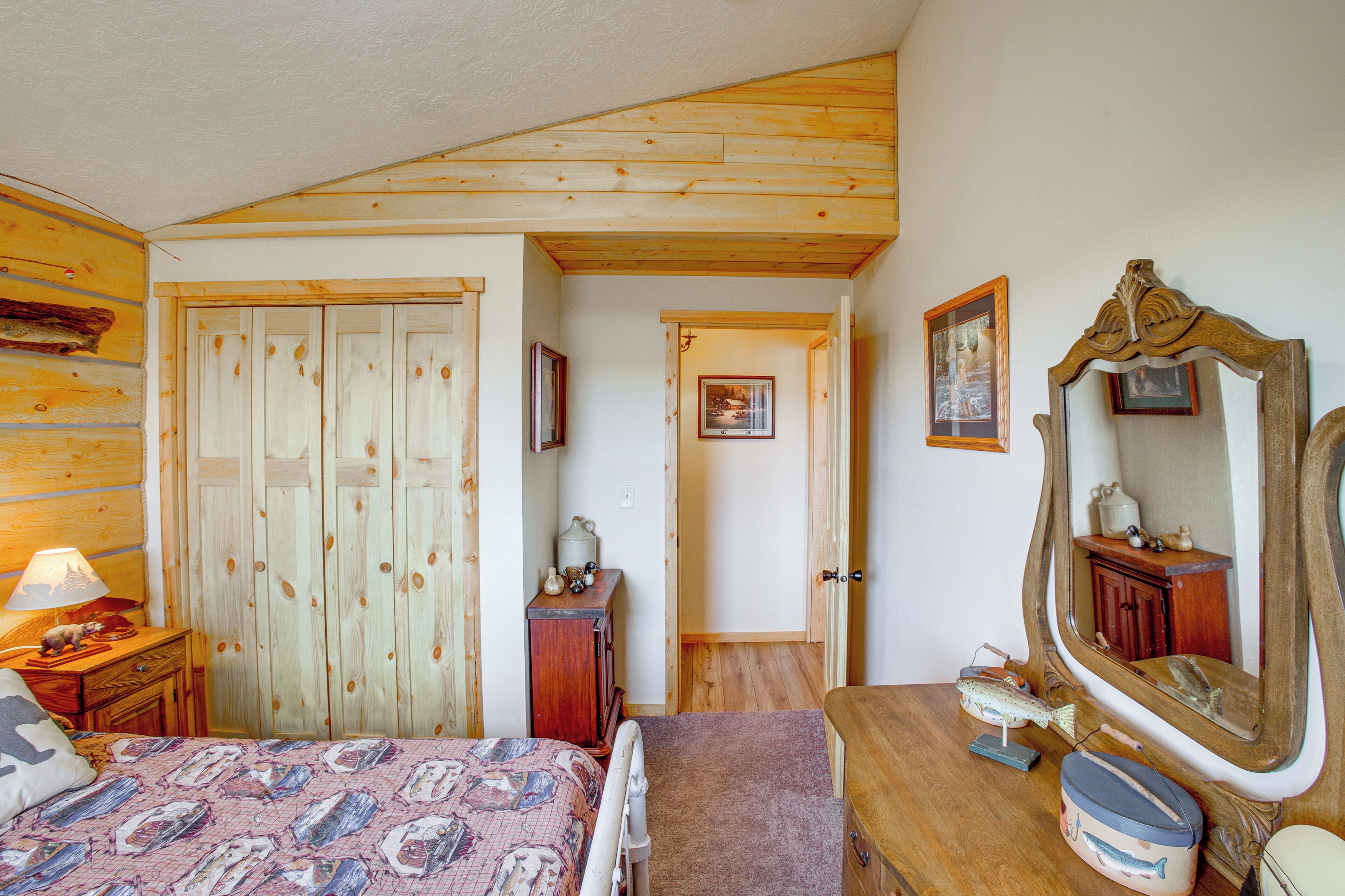 Un dormitorio con paredes de madera, una cama con colcha estampada, un aparador, un espejo y una puerta que da a otra habitación.
