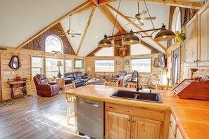 A spacious log cabin interior with a kitchen island, dining area, and a large window overlooking a scenic landscape. - Roberts Home w/ Western Decor & Mountain Views! (Roberts)