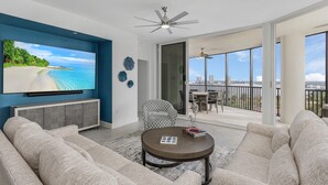 A modern living room with a large TV, comfortable seating, and a balcony with a city view. - Grandview #603 - 4142 Bay Beach Ln 3 Bedroom Condo by RedAwning (Fort Myers Beach)