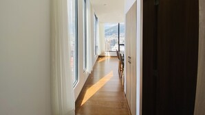 Un largo y estrecho pasillo con amplios ventanales en un lado, suelo de madera y vistas a un paisaje urbano y montañas al fondo.