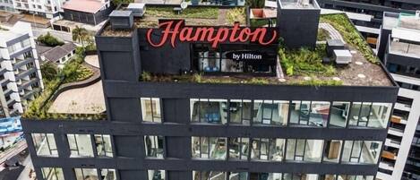 A hotel building with a rooftop garden and a large sign reading "Hampton by Hilton".