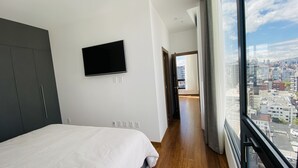 Un dormitorio moderno con una cama grande, un televisor montado y vistas a la ciudad a través de un ventanal amplio.