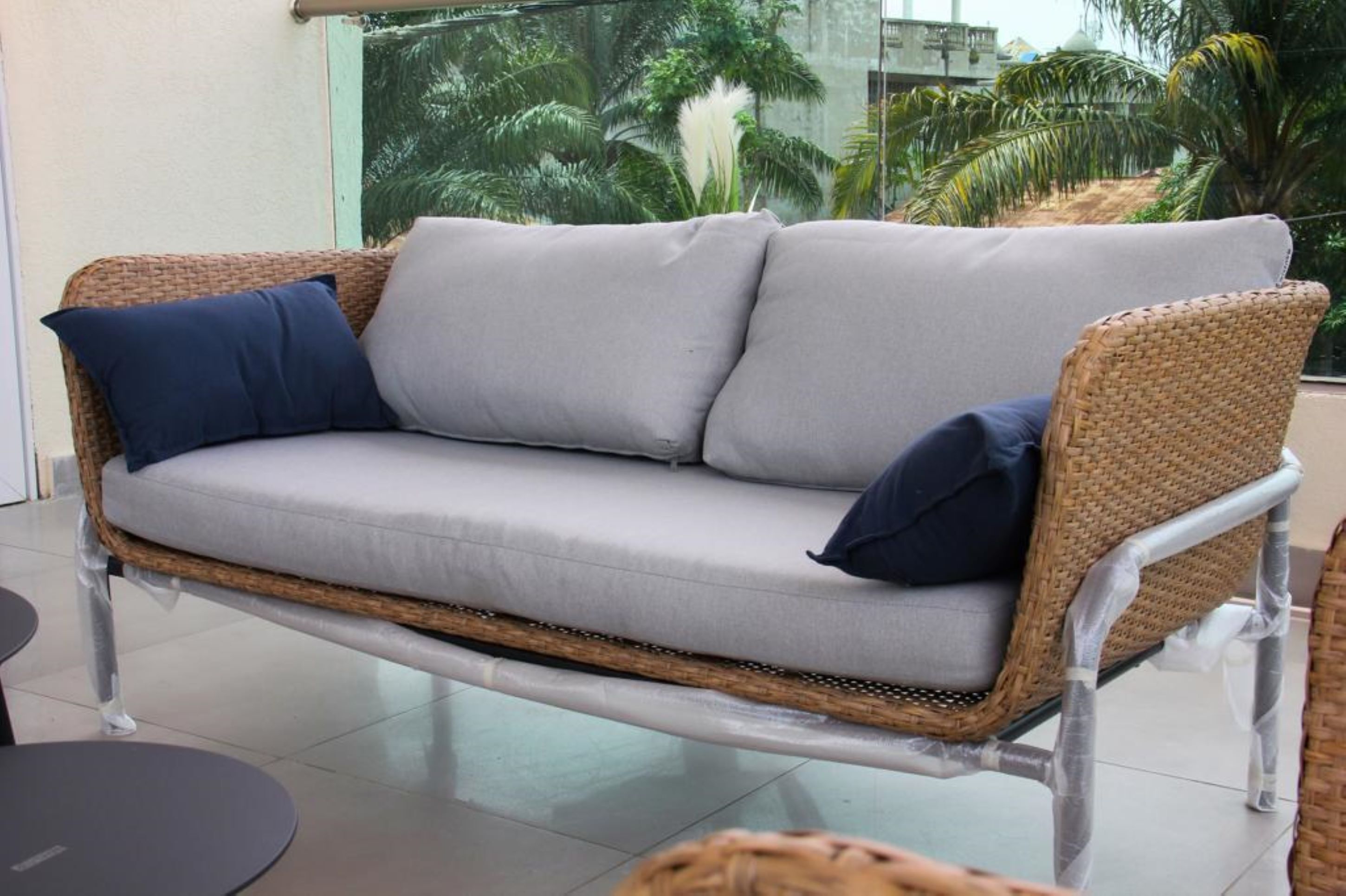 A wicker sofa with grey cushions and a dark blue pillow, positioned indoors with a view of palm trees through a glass window.