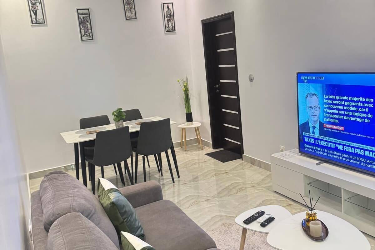 A modern living room with a grey sofa, a dining area, and a flat-screen TV displaying a news channel.
