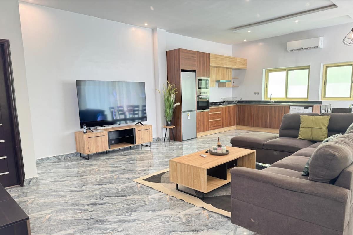 A modern living room with a flat-screen TV, a sofa, a coffee table, and a kitchen area with wooden cabinets and a refrigerator.
